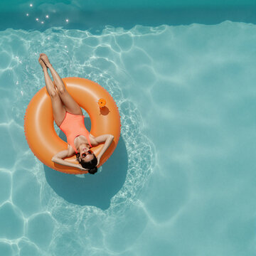 relaxed young woman in orange swimsuit lounging on pool float under sunlight in tranquil summer pool