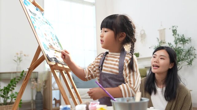 Young child painting at easel with brush, apron, watercolor paint in bright art studio, girl creating canvas artwork near helper, plant, calm home creativity