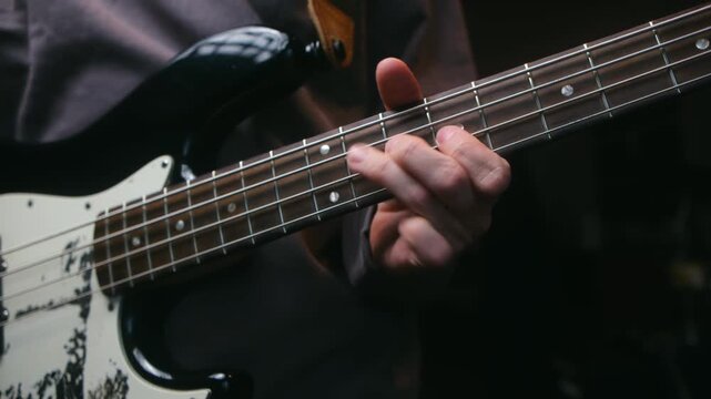 Close up skilled bass player hand fretting notes on neck of black and white electric bass guitar, studio recording session in dark environment. Musician playing vintage bass guitar fretboard