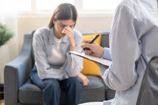 Female psychologist taking notes on clipboard during therapy session with a depressed Asian woman patient