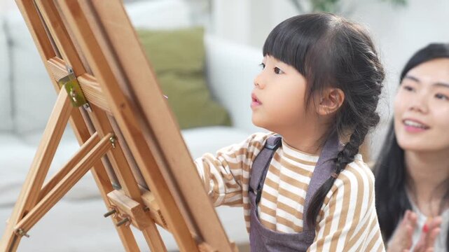 Child girl mother painting easel brush art home family learning Asian creative education canvas indoor lesson hobby striped shirt braid focus paintbrush bright room expression