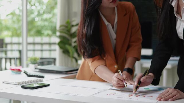 Two female coworkers standing at a desk and reviewing business documents together, pointing at printed charts during a collaborative planning and analysis session in the office.
