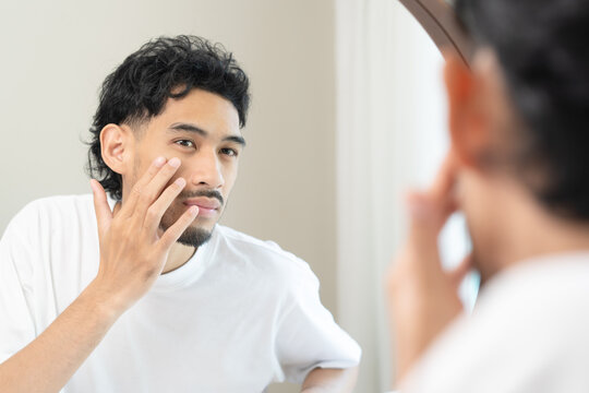 Handsome young Asian man looking in the mirror and checking his skin during morning routine at home