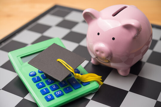 Graduation cap, calculator, piggy bank on chessboard. Personal financial management for education, repayment student loan or debt, tuition, find out scholarship for studying abroad and so on concept.