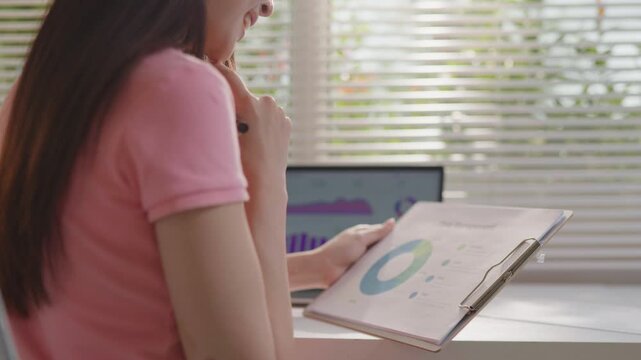 Woman working on a laptop while reviewing printed business charts and graphs, analyzing data and performance reports in a bright home office workspace.