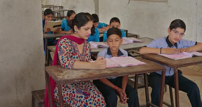 Indian young adult female tutor sitting on bench talk speak do helping to study child boy indoor class room. Teen student wear uniform hold pencil look book read exam note at rural area village place