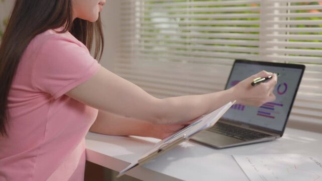 Woman working on a laptop while reviewing printed business charts and graphs, analyzing data and performance reports in a bright home office workspace.