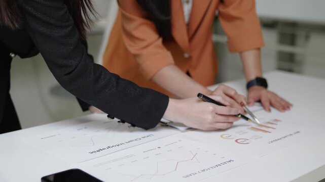 Close-up of two businesswomen discussing printed charts and reports on a desk, pointing at data together during a brainstorming and business analysis session.