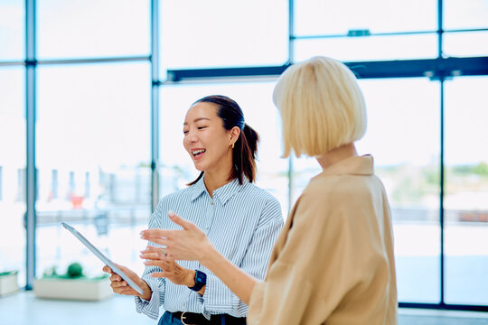 Two diverse businesswomen discussing business ideas, collaborating and smiling during a brainstorming meeting with a digital tablet