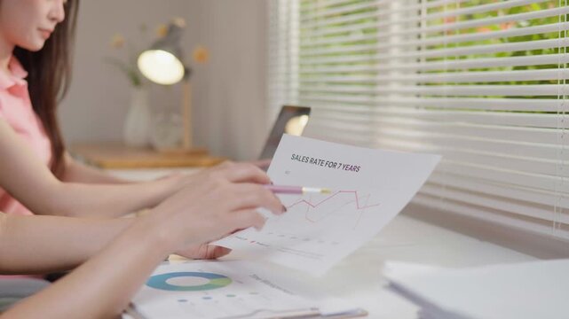 Close-up of a woman&rsquo;s hand pointing at a printed business graph and chart report on a desk, with a laptop in the background, representing data analysis, financial review, and office planning.