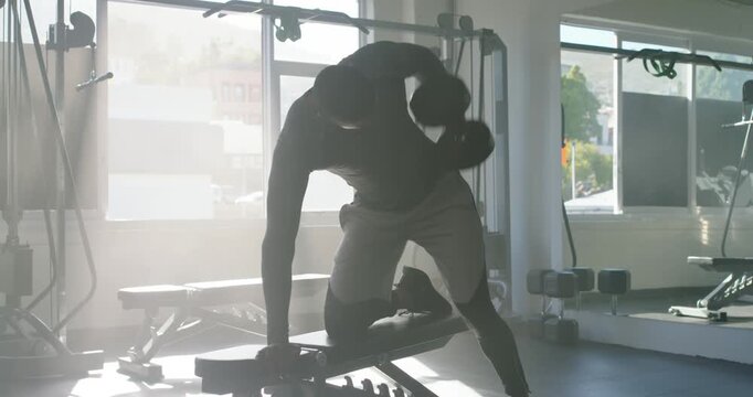 African adult man in longsleeve and shorts grabbing dumbbell doing rows for strength at gym bench