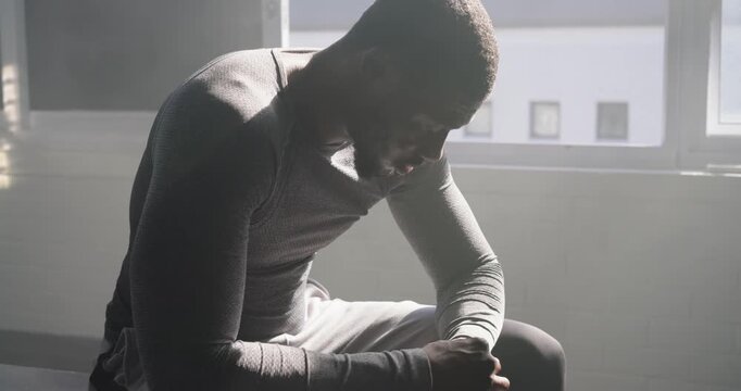 African American man sitting on gym bench by window, breathing and gathering strength, lifting head