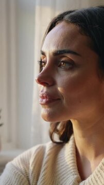 Vertical video: Gazing woman in cream sweater by window with plant, light drawing blink, soft smile