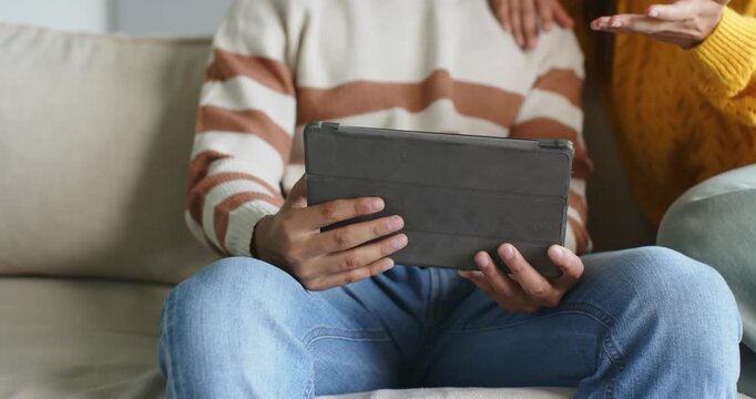 African American couple sitting on light sofa, woman gesturing at folio tablet causing man holding