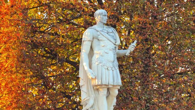 Classical white marble statue standing against a rich backdrop of golden and bronze autumn tree leaves in the Tuileries Garden in Paris, France