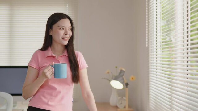 Young woman holding a blue coffee mug at home, enjoying a quiet moment by the window in a bright and cozy interior.