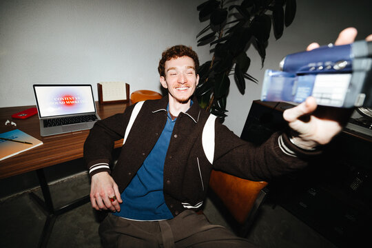 The camera captures a smiling man making video content at his laptop desk.