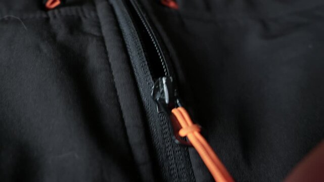 Close-up of fingers pulling a bright orange zipper cord on a black technical jacket. Concept of outdoor gear preparation, staying warm, and getting ready for a winter hike.