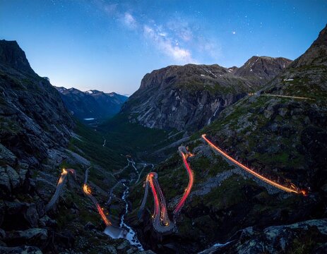 Long exposure nightscape of Trollstigen switchbacks in Norway with flowing red light trails and Stigfossen waterfall under a majestic arched Milky Way galaxy starscape. 8k.