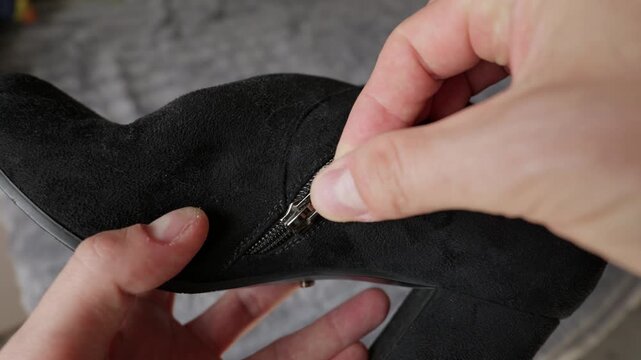 Close-up of a person hands examining the silver zipper on a black suede ankle boot. The boot features a chunky block heel and soft textured material. Soft light highlights the metal teeth.