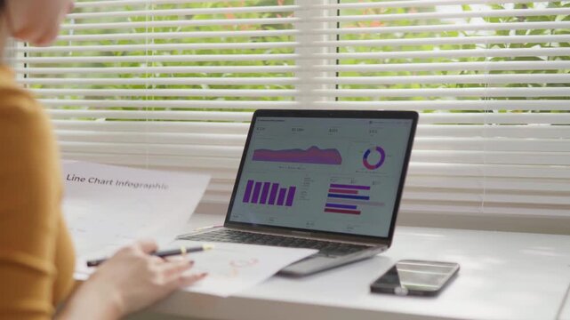 Woman working on a laptop with charts and graphs on screen, reviewing documents and analyzing business data at a home office desk.