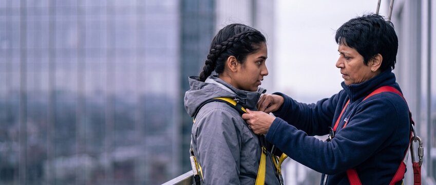 Industrial climber safety gear rooftop inspection with female instructor preparing young woman for high altitude work on glass skyscraper ledge, team trust and courage concept portrait outdoor scene.