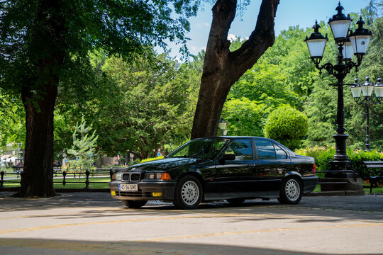Three-quarters profile view of a vintage black BMW 3 Series (E36) sedan parked in a green shaded park area near a classic street lamp post in Subotica, Serbia, on June 7, 2026
