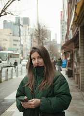 Fototapeta premium Young woman in green puffer jacket holding smartphone smiling on a Tokyo street with traditional shop signage and city buildings in background 