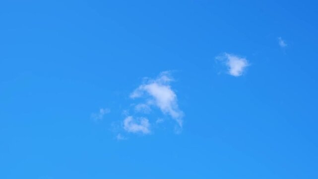 A clear bright blue sky with a few small, wispy white clouds scattered across it. Panoramic sunlit vast skyscape. Vibrant cloudscape. High quality 4k footage.