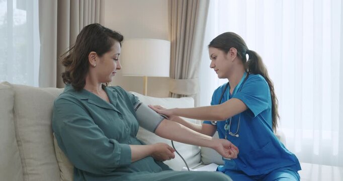 Nurse Taking Blood Pressure of Female Patient at Home, Home Care Visit with Healthcare Worker in Blue Scrubs Using Blood Pressure Cuff on Sofa in Living Room