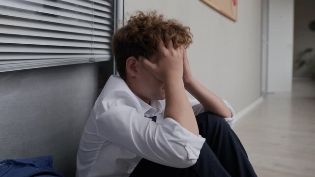 Caucasian teenager boy sitting on school corridor floor, covering face with hands, crying and showing distress. Visuals represent student mental health and anti-bullying campaigns