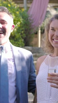 Vertical video: Toasting couple in gray suit, bride raising flutes in yard as guests join wedding