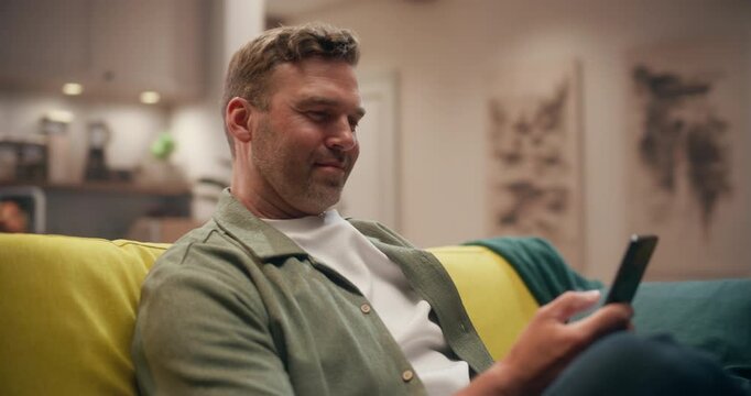 360 Degree. Caucasian Man Sits on Couch at Home, Using a Smartphone to Shop Online, Make Ecommerce Purchases, and Browse the Internet in a Cozy Living Room. Cinematic Arc Tracking Shot.