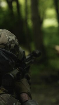 Camouflaged soldier in the middle of a dark forest on a recon mission. Aiming a AR-15 carbine rifle with a red collimator and a magnifier. Military training