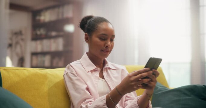 360 Degree. Black Woman Sits on a Living Room Couch Using a Smartphone to Browse Online Stores, Shop, Make Mobile Purchases, Check Social Media, Work Remotely From Home. Cinematic Arc Tracking Shot.