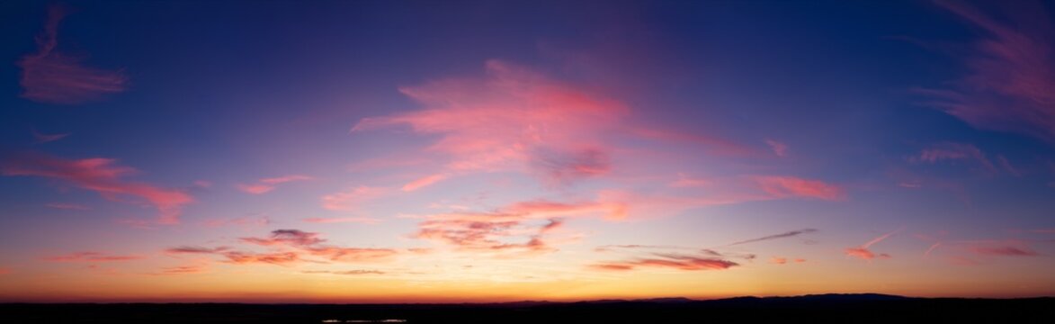 Panoramic drone view of a colorful sunset sky with soft pink cloud streaks, warm glow near the horizon and a clean dark landscape silhouette, ideal for background use. Colors: pink, blue, orange.
