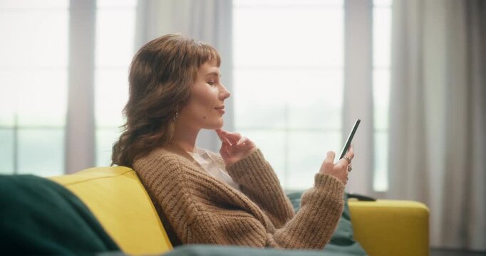 360 Degree. Caucasian Woman Sits Comfortably on a Living Room Couch Using a Smartphone to Browse Social Media Feeds, Shop Online Stores, Reply to Messages, and Work From Home. Cinematic Tracking Shot.