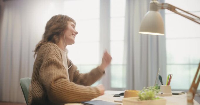 361 Degree. Young Caucasian Woman Listening to Music With Earphones While Using Laptop Computer at Home Desk, Dancing Joyfully, Browsing Internet and Remote Work in Apartment. Cinematic Tracking Shot.