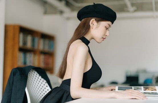 Young Asian Woman in Black Sleeveless Top and Beret Sitting at Office Desk Side Profile Reading Magazine in Modern Creative Studio Interior Lifestyle Fashion Portrait Brunette Korean Beauty