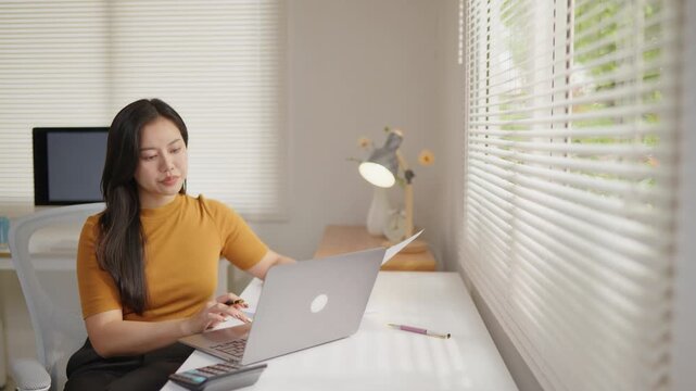 Woman working on a laptop at a home office while reviewing documents, ideal for remote work, online business, administration, planning, productivity and workflow concepts.