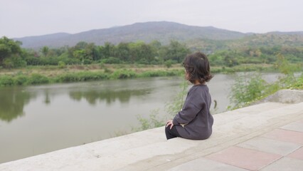 Fototapeta premium A young toddler sits alone on a concrete embankment, quietly observing the river and mountains, embodying a peaceful moment of childhood contemplation.