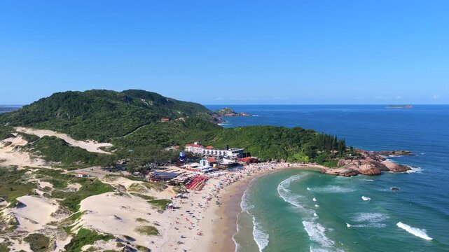 Aerial view of Joaquina Beach with turquoise waves, sand dunes, and rocky coastline in Florian&oacute;polis.