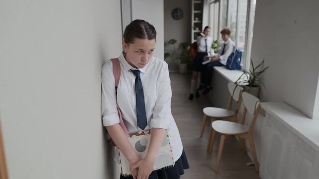 Lonely Caucasian teenage girl leaning against wall in school corridor while classmates talking in background. Ideal for videos addressing teenage mental health, school bullying, and social isolation
