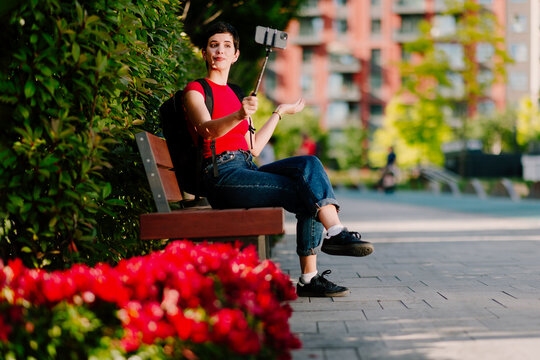Young Caucasian woman sitting on a park bench while recording online content with a smartphone mounted on a selfie stick.