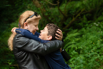 Fototapeta premium A mother lovingly embraces her smiling son during a walk in lush green forest. Sweet moments of communication between a mother and her teenage son