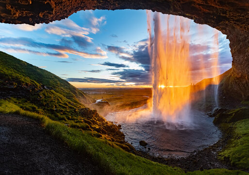 Impressive waterfall &ldquo;Seljalandsfoss&ldquo; in Iceland seen from a cave behind the sunlit water curtain at midnight in June. Colorful sunset panorama in wild nature of volcanic scenery. Major attraction.