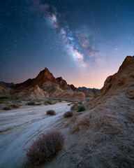 Obraz premium Milky Way arches above rugged desert peaks. Soft twilight paints sky with hues of rose and blue. Distant stars glitter like scattered diamonds. Dry earth and sparse shrubs frame the scene