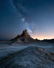 Fototapeta premium Dark desert peak rises beneath swirling Milky Way. Starry sky glows softly above rocky horizon. Dusk paints horizon with warm, fading light. Silent, ancient land holds timeless cosmic wonder