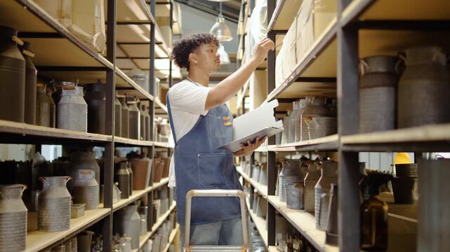 Focused young male small business owner in a denim apron checks inventory on a clipboard in a rustic warehouse. He inspects stock on metal shelves filled with vintage pitchers and shipping boxes.