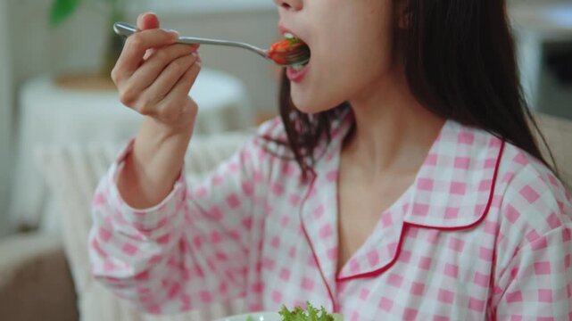Healthy Asian woman in pajamas eating fresh green salad for breakfast on sofa, healthy eating and diet lifestyle concept.